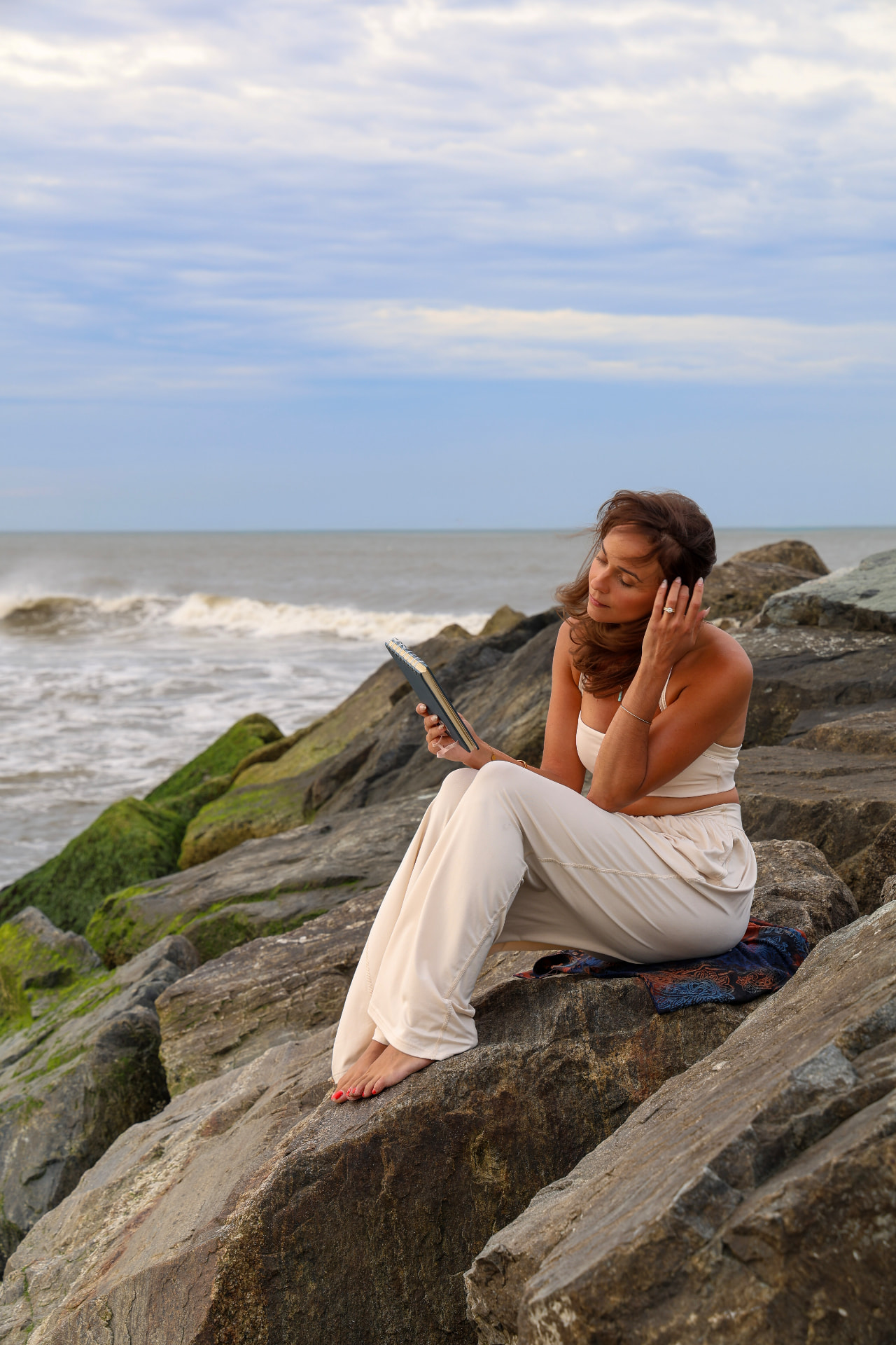 Nicole sitting on ocean rocks with her journal, wearing white